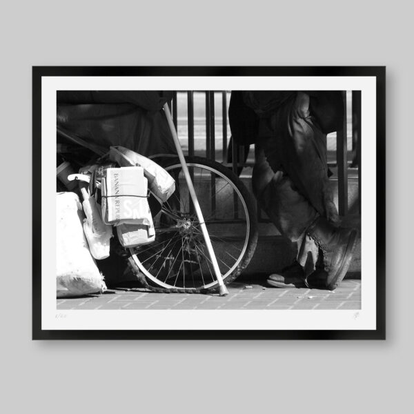 Fotografía artística en blanco y negro. Titulo: Let's roll. Se ve parte de las botas de un hombre al lado de una rueda de su bicicletas. Hay diarios y revistas que están amarrados.